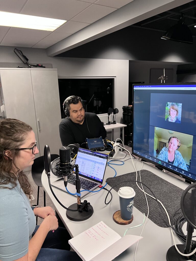 People in a podcast recording studio, with a video call on screen. Transcribed Text: The notebook has handwritten text, partially visible, in red ink.