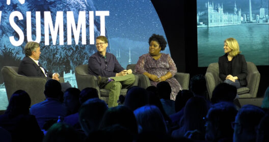 Four panelists seated in armchairs on a stage at a summit, with the word "SUMMIT" and nighttime cityscape on the screen behind them, in front of an audience.