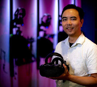 A smiling man holding a VR headset in front of him with camera equipment in the softly lit, colorful background.