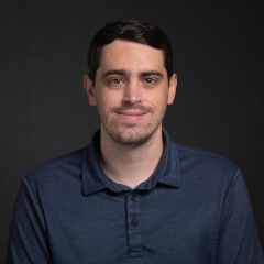 Portrait of a man with short dark hair, wearing a navy-blue polo shirt, against a dark background.