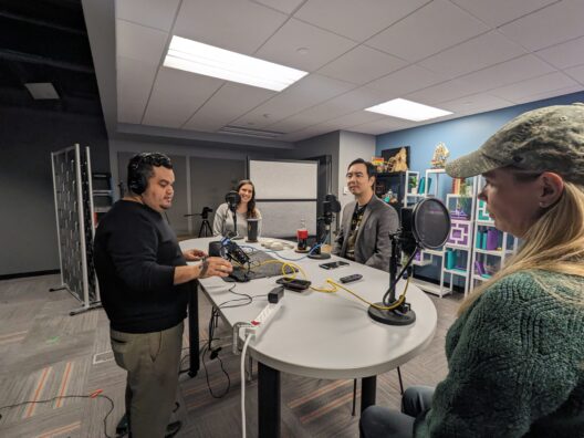 Four people in a podcast recording session with microphones and audio equipment on a white round table in a modern, decorated room.